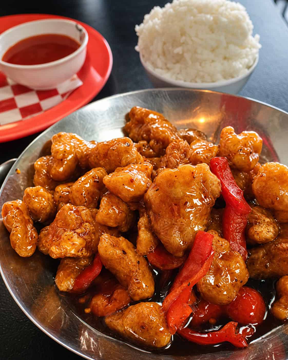 A steel wok bowl of Bamboo House Orange Chicken — crisp golden chicken in a glossy orange sauce with strips of red pepper — beside a bowl of steamed white rice and a cup of sauce.