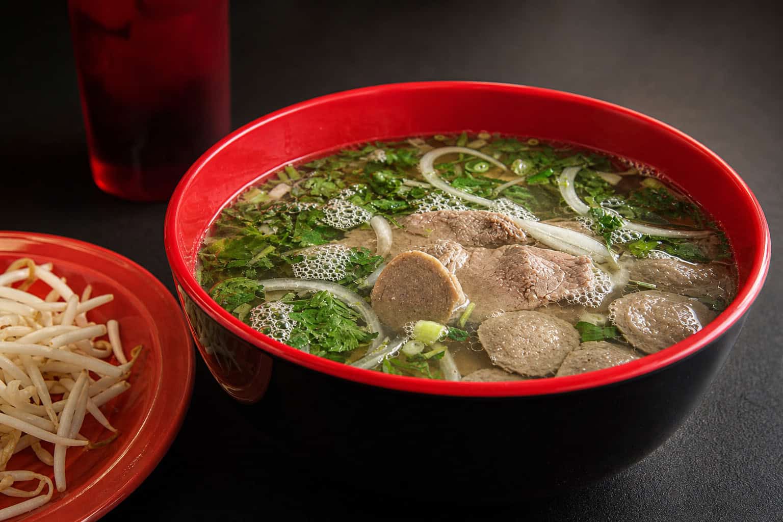 A red bowl of Bamboo House rare beef and meatball pho with cilantro, sliced onion, and noodles in clear broth, served beside a plate of bean sprouts.